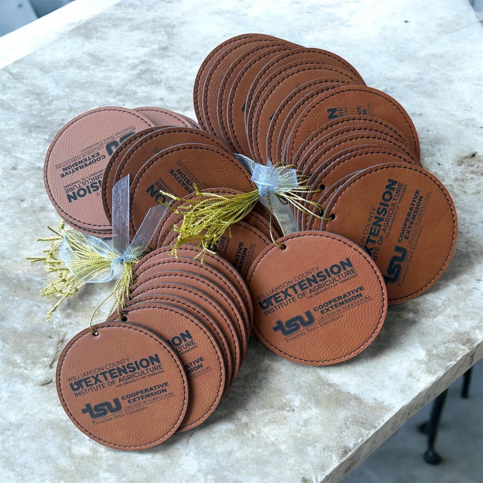 Round brown leatherette Christmas ornaments engraved with sample logos, stacked with gold tassels